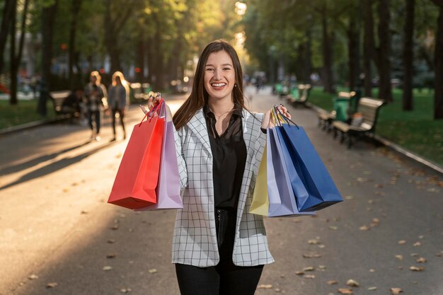 Mujer caucásica joven hermosa de moda que lleva bolsas de compras coloridas en el callejón del fondo del parque