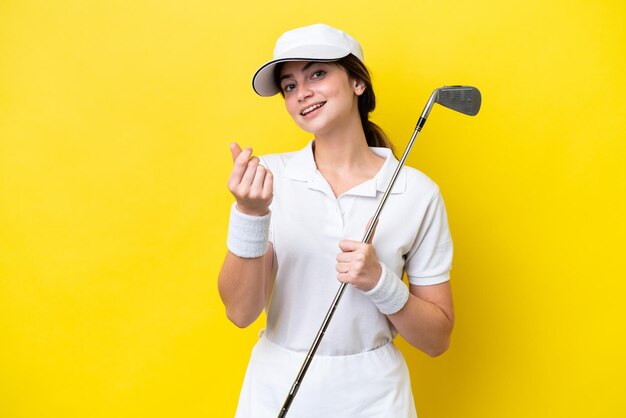 Mujer caucásica joven jugando al golf aislado sobre fondo amarillo haciendo gesto de dinero