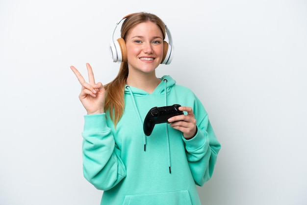 Mujer caucásica joven jugando con un controlador de videojuegos aislado sobre fondo blanco sonriendo y mostrando el signo de la victoria