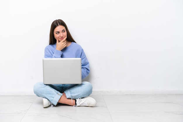 Mujer caucásica joven con una laptop sentada en el suelo aislada de fondo blanco que tiene dudas y con una expresión facial confusa