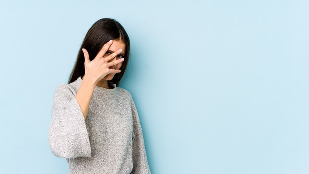 La mujer caucásica joven en la pared azul parpadea a la cámara con los dedos, avergonzada que cubre la cara.