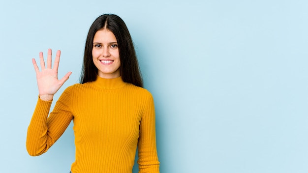 Mujer caucásica joven en la pared azul que sonríe alegre que muestra el número cinco con los dedos.