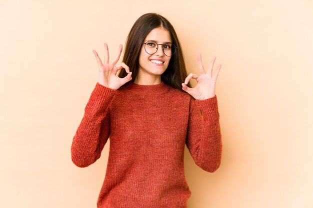 Mujer caucásica joven en la pared beige alegre y confiada que muestra gesto aceptable.