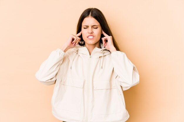 Mujer caucásica joven en la pared beige que cubre las orejas con los dedos, estresado y desesperado por un ambiente ruidosamente.