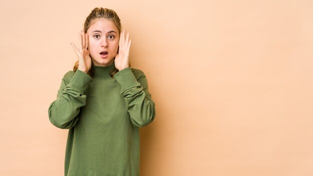 Mujer caucásica joven en la pared beige sorprendida y conmocionada.