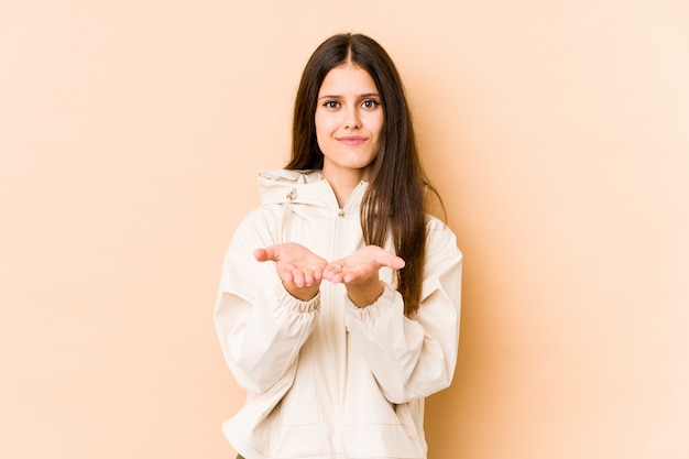 Mujer caucásica joven en la pared beige sosteniendo algo con las palmas, ofreciendo a la cámara.