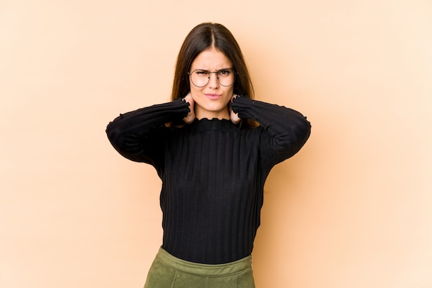 Mujer caucásica joven en la pared de color beige que sufre dolor de cuello debido al estilo de vida sedentario.