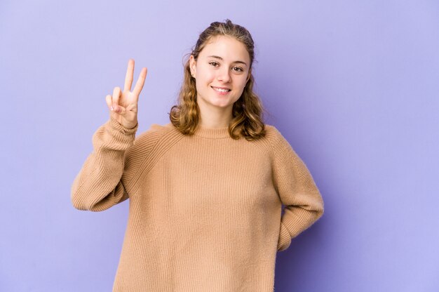 Mujer caucásica joven en la pared púrpura que muestra la muestra de la victoria y que sonríe ampliamente.