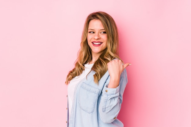 La mujer caucásica joven en pared rosa señala con el dedo pulgar lejos, riendo y despreocupado.