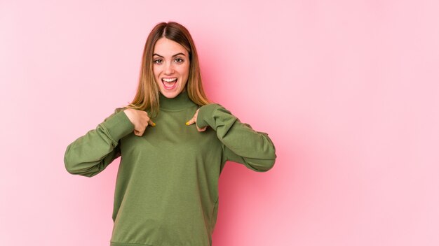 La mujer caucásica joven en la pared rosada sorprendió señalar con el dedo, sonriendo ampliamente.