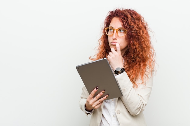 Mujer caucásica joven pelirroja de negocios sosteniendo una tableta relajada pensando en algo mirando un espacio de copia