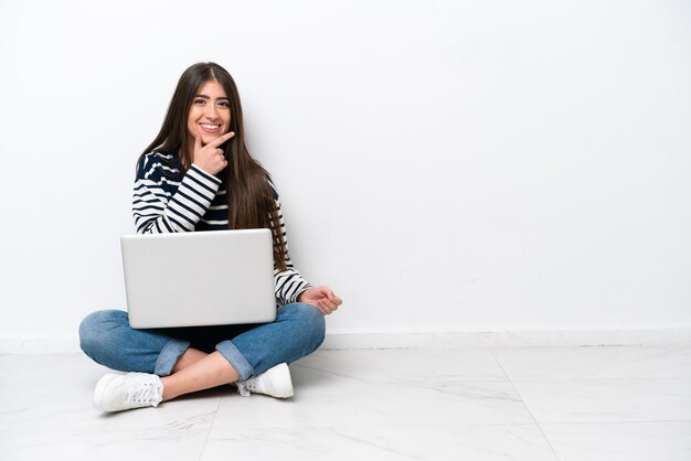 Mujer caucásica joven con un portátil sentado en el suelo aislado sobre fondo blanco sonriendo