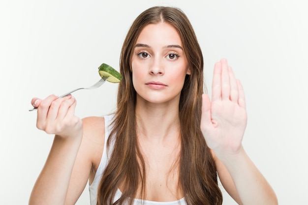 Mujer caucásica joven que come el pepino que se coloca con la mano extendida que muestra la señal de stop, previniéndole.