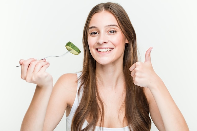 Mujer caucásica joven que come el pepino que sonríe y que levanta el pulgar para arriba