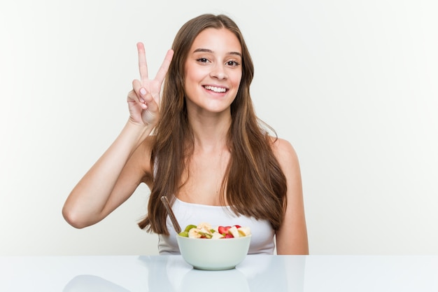 Mujer caucásica joven que come el tazón de fuente de fruta que muestra la muestra de la victoria y que sonríe ampliamente.