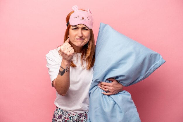 Mujer caucásica joven que lleva un pijama que sostiene la almohada aislada en el fondo rosado que muestra el puño a la cámara expresión facial agresiva