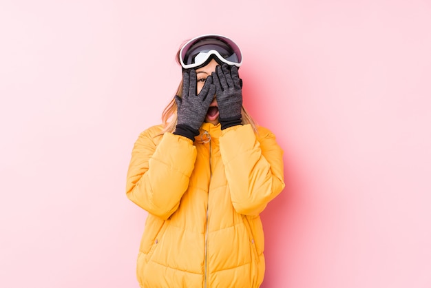 La mujer caucásica joven que lleva una ropa de esquí en una pared rosada parpadea entre los dedos asustada y nerviosa.