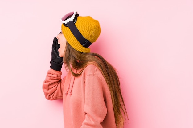 La mujer caucásica joven que llevaba un esquí viste aislado gritando y sosteniendo la palma cerca de boca abierta.