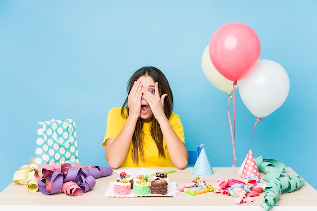 Mujer caucásica joven que organiza un parpadeo de cumpleaños a través de los dedos asustados y nerviosos.