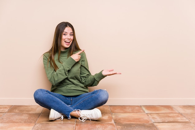 La mujer caucásica joven que se sentaba en el piso aisló excitado sosteniendo un espacio de la copia en la palma.