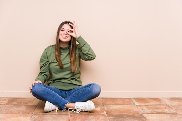 La mujer caucásica joven que se sentaba en el piso excitó guardar gesto aceptable en ojo.