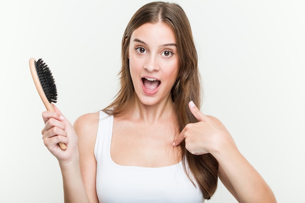 La mujer caucásica joven que sostenía un cepillo de pelo sorprendió señalando a sí mismo, sonriendo ampliamente.