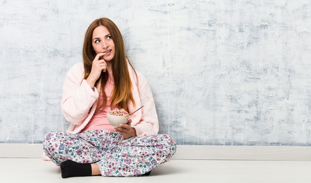 La mujer caucásica joven que sostenía un tazón de cereal relajado pensando en algo que miraba un espacio de la copia.