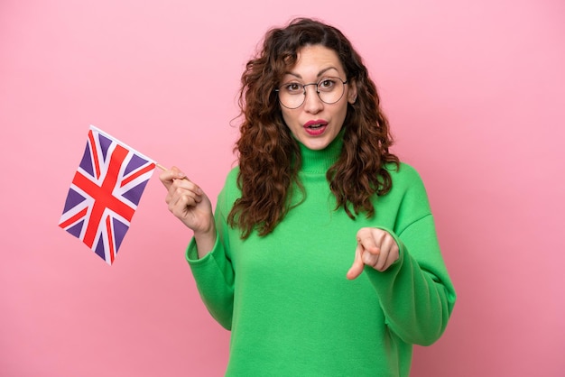 Mujer caucásica joven que sostiene la bandera inglesa aislada en el fondo rosado sorprendida y que señala al frente
