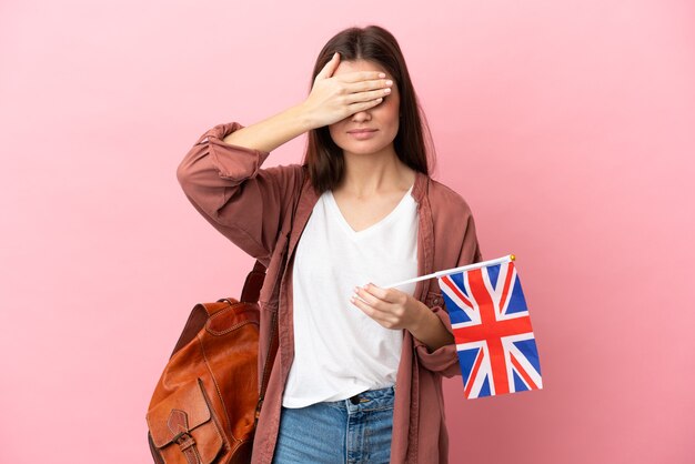 Mujer caucásica joven que sostiene una bandera del Reino Unido aislada en la pared rosada que cubre los ojos con las manos. No quiero ver algo