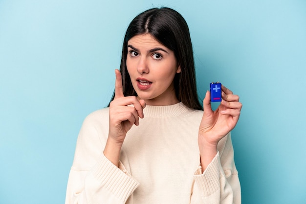 Mujer caucásica joven que sostiene una batería aislada en el fondo azul que tiene un concepto de inspiración de idea