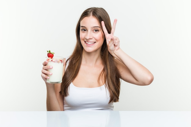 Mujer caucásica joven que sostiene un batido que muestra el signo de la victoria y que sonríe ampliamente.