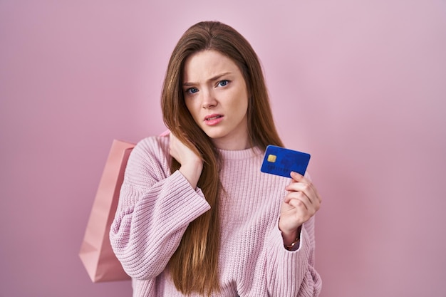 Mujer caucásica joven que sostiene el bolso de compras y la expresión despistada y confusa de la tarjeta de crédito. concepto de duda.