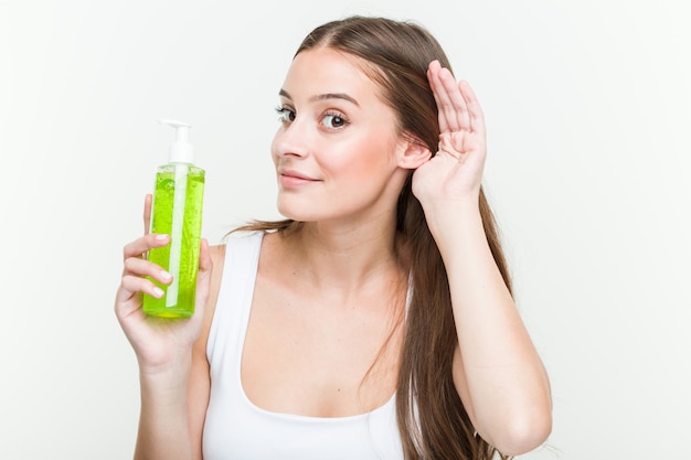 Mujer caucásica joven que sostiene una botella de aloe vera tratando de escuchar un chisme.