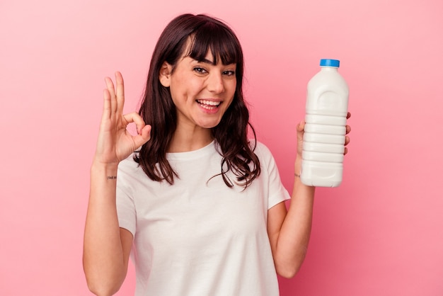 Mujer caucásica joven que sostiene una botella de leche aislada en la pared rosada alegre y confiada que muestra el gesto aceptable