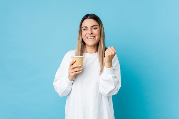 Mujer caucásica joven que sostiene un café para llevar que anima despreocupado y emocionado. Concepto de victoria