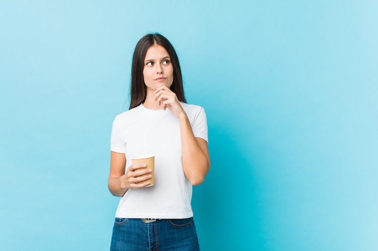 Foto mujer caucásica joven que sostiene un café para llevar que mira de lado con expresión dudosa y escéptica.