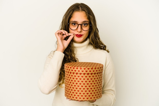 Mujer caucásica joven que sostiene una caja del día de San Valentín del corazón aislada con los dedos en los labios que guardan un secreto.