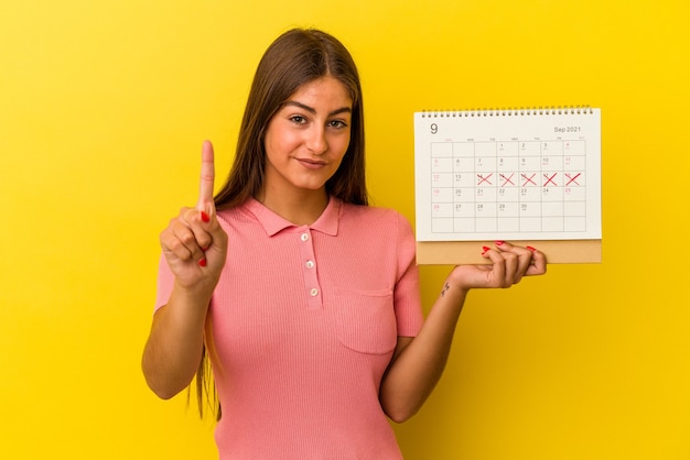 Mujer caucásica joven que sostiene un calendario aislado en fondo amarillo que muestra el número uno con el dedo.