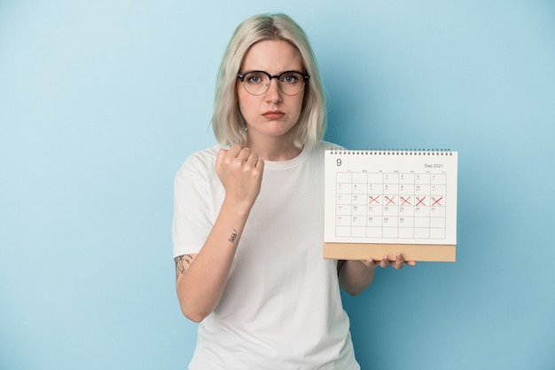 Mujer caucásica joven que sostiene el calendario aislado en el fondo azul que muestra el puño a la cámara, expresión facial agresiva.