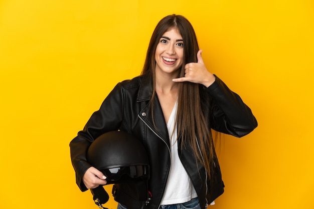 Mujer caucásica joven que sostiene un casco de la motocicleta aislado en la pared amarilla que hace el gesto del teléfono. Llámame señal