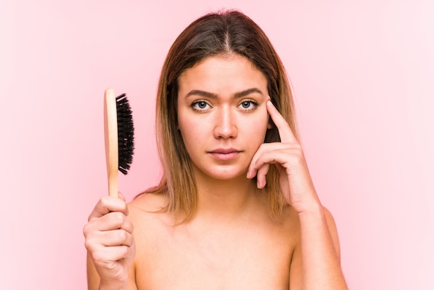 Mujer caucásica joven que sostiene un cepillo para el pelo aislado señalando su sien con el dedo, pensando, centrado en una tarea.