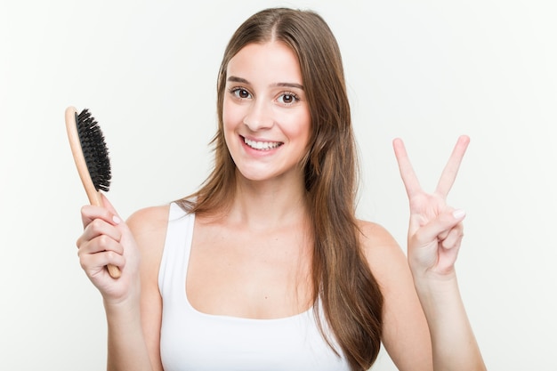 Mujer caucásica joven que sostiene un cepillo de pelo que muestra el número dos con los dedos.