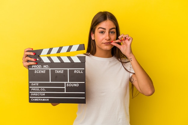 Mujer caucásica joven que sostiene la claqueta aislada en el fondo blanco con los dedos en los labios que guardan un secreto.