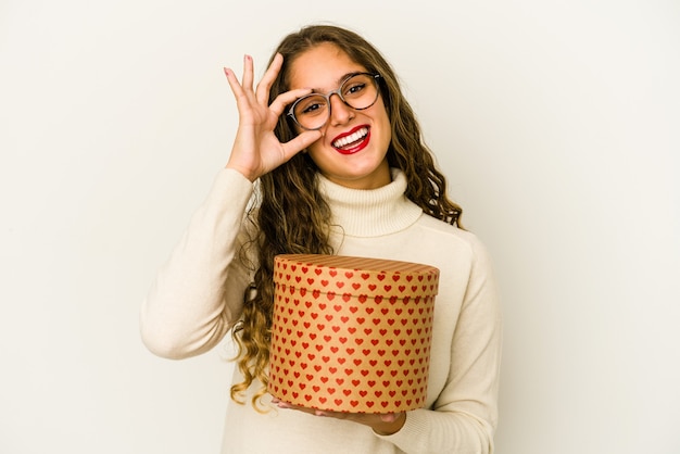 Mujer caucásica joven que sostiene un cuadro del día de tarjetas del día de San Valentín del corazón aislado emocionado manteniendo el gesto aceptable en el ojo.