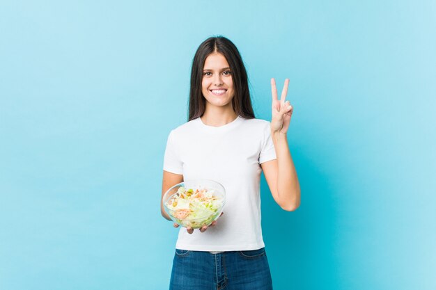 Mujer caucásica joven que sostiene una ensalada que muestra el número dos con los dedos.