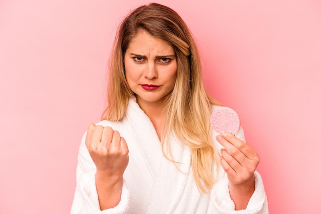 Mujer caucásica joven que sostiene la esponja facial aislada en el fondo rosado que muestra el puño a la cámara expresión facial agresiva