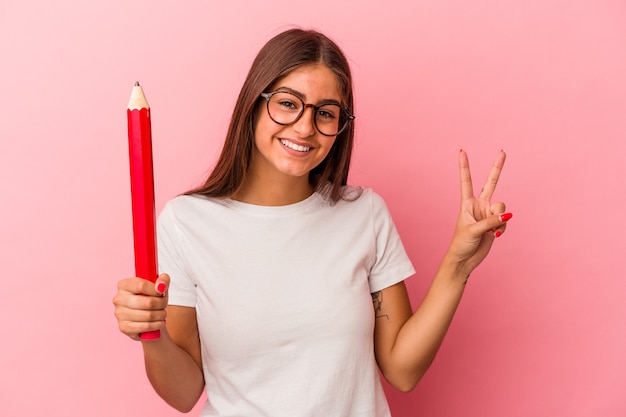 Mujer caucásica joven que sostiene un lápiz grande aislado en fondo rosado que muestra el número dos con los dedos.