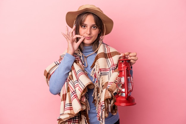 Mujer caucásica joven que sostiene la linterna del vintage aislada en fondo rosado con los dedos en los labios que guardan un secreto.