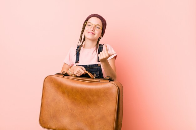 La mujer caucásica joven que sostiene una maleta que señala con el dedo en usted como invitando se acerca.