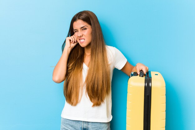 Mujer caucásica joven que sostiene una maleta de viaje que cubre las orejas con las manos.